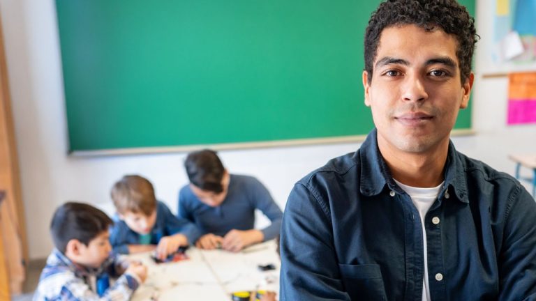 Professor jovem em destaque, de braços cruzados, olhando diretamente para a câmera em uma sala de aula. Ao fundo, um grupo de crianças está sentado em uma mesa, concentrado em uma atividade manual. Ao fundo há um grande quadro verde e paredes com cartazes coloridos parcialmente visíveis. O foco está no educador, transmitindo segurança e protagonismo.