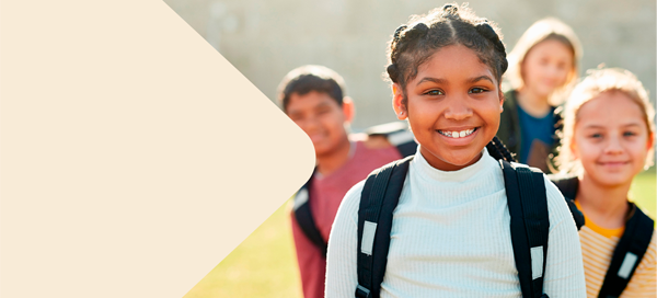 Grupo de crianças com mochilas sorri ao ar livre em um dia ensolarado. Em destaque, uma menina negra de cabelos trançados usa blusa clara e sorri para a câmera.