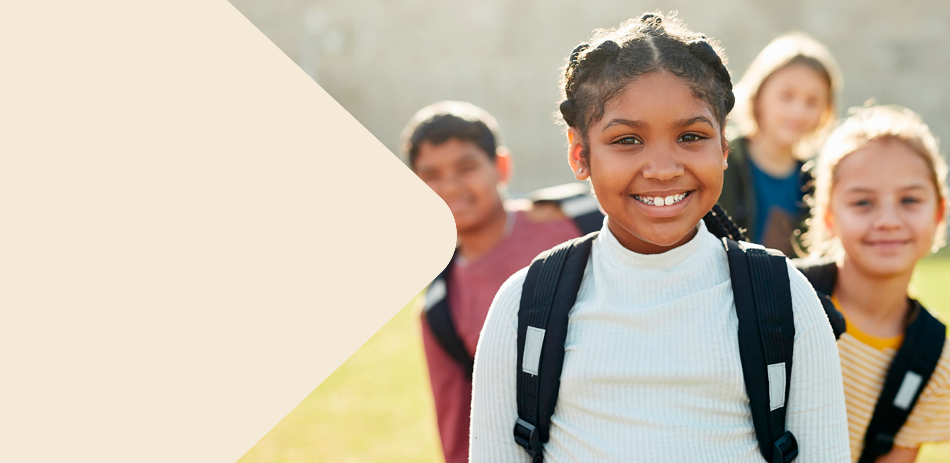 Grupo de crianças com mochilas sorri ao ar livre em um dia ensolarado. Em destaque, uma menina negra de cabelos trançados usa blusa clara e sorri para a câmera.