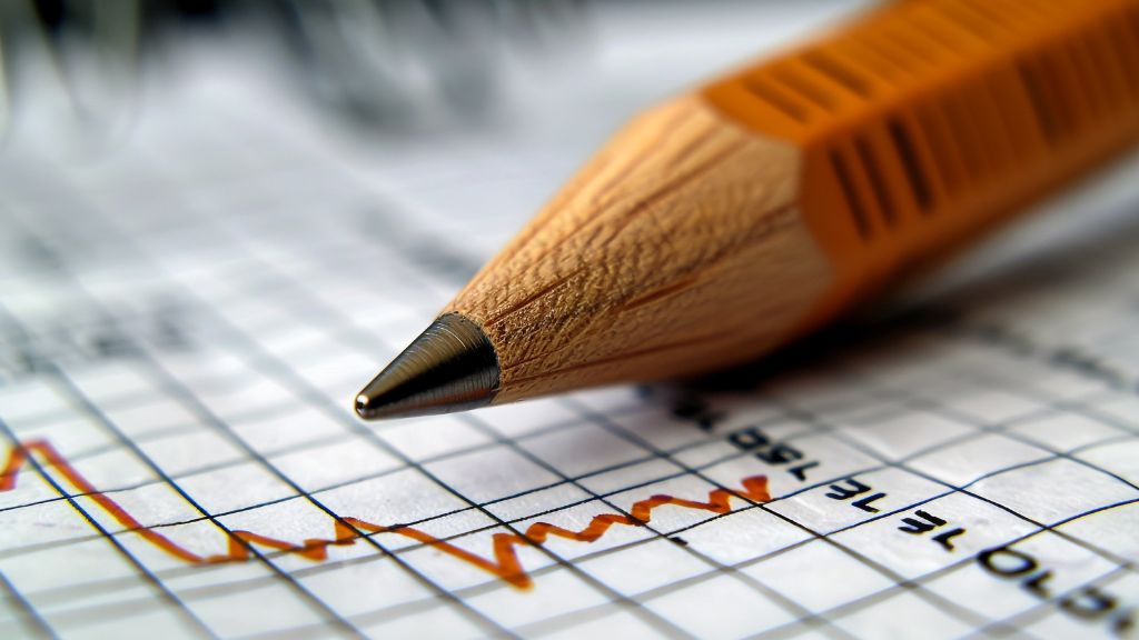 Lápis laranja apontado de perto sobre uma folha quadriculada com gráfico de linha em vermelho, indicando variações de dados. A imagem remete à análise estatística ou estudo de dados educacionais.