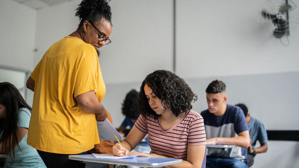 Professora de blusa amarela orienta uma aluna durante atividade em sala de aula. A estudante escreve atenta, enquanto outros alunos, ao fundo, também realizam suas tarefas. A sala tem paredes claras e iluminação natural, criando um ambiente de concentração e aprendizado.