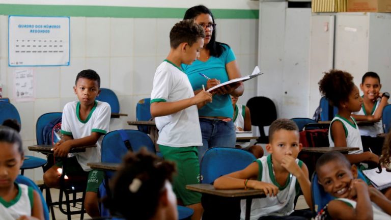 Crianças em sala de aula interagem entre si e com a professora. Os alunos usam uniforme escolar branco com verde e estão sentados em carteiras azuis. Uma professora ajuda um estudante em pé, segurando um caderno. O ambiente é claro e organizado, com cartazes educativos na parede.