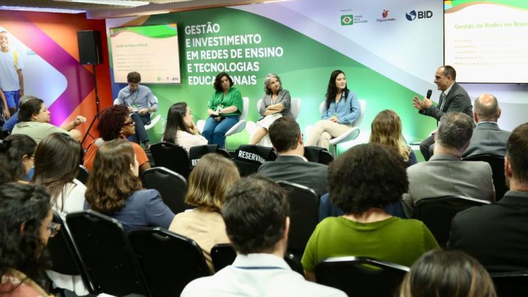 A imagem mostra um evento ou seminário sobre educação, com o tema “Gestão e Investimento em Redes de Ensino e Tecnologias Educacionais”, conforme destacado no painel de fundo. À frente, um painel com cinco pessoas está reunido para um debate ou apresentação. Um dos participantes, à direita, fala ao microfone enquanto os demais escutam atentamente. A plateia está composta por dezenas de pessoas, sentadas e concentradas na discussão. No canto superior direito da imagem, estão visíveis os logotipos do Todos Pela Educação, BID (Banco Interamericano de Desenvolvimento) e Segurança Pública.