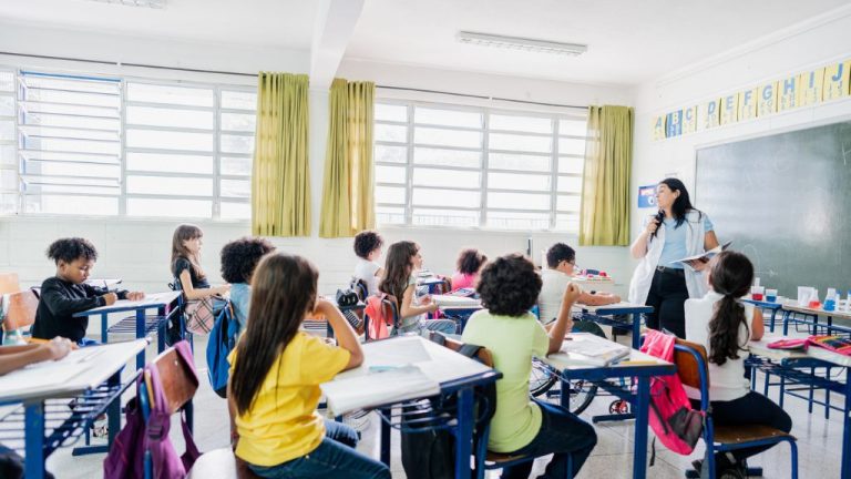 Sala de aula com crianças sentadas em carteiras azuis, prestando atenção à professora que fala em frente ao quadro negro. O ambiente é iluminado por janelas amplas com cortinas verdes, e há materiais escolares sobre as mesas.