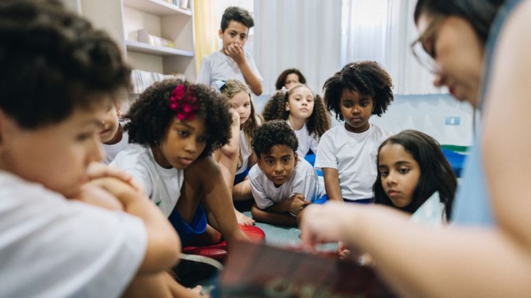 Um grupo de crianças, vestindo uniformes escolares brancos e azuis, está sentado em roda, atento ao livro que a professora lê. Elas parecem concentradas e curiosas. O ambiente é uma sala de aula ou biblioteca, com prateleiras de livros ao fundo e iluminação natural.