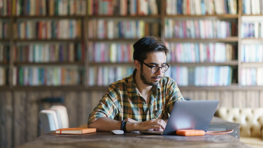 A imagem mostra um homem jovem sentado em uma biblioteca, concentrado enquanto usa um notebook. Ele tem pele clara, barba curta e usa óculos de grau. Está vestindo uma camisa xadrez em tons de verde, amarelo e marrom. Sobre a mesa há um caderno laranja e um disco rígido externo conectado ao computador. Ao fundo, há prateleiras de madeira cheias de livros coloridos, compondo um ambiente acolhedor e de estudo.