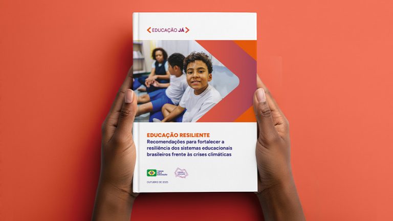 A imagem mostra um par de mãos segurando um relatório impresso com capa branca. Na capa há uma foto de um garoto sorrindo em sala de aula, acompanhada de um gráfico em forma de seta nas cores laranja e roxa. O título do documento é “Educação Resiliente – Recomendações para fortalecer a resiliência dos sistemas educacionais brasileiros frente às crises climáticas”. A publicação é assinada pelo Todos Pela Educação e pelo movimento Vozes da Educação, com data de outubro de 2025.