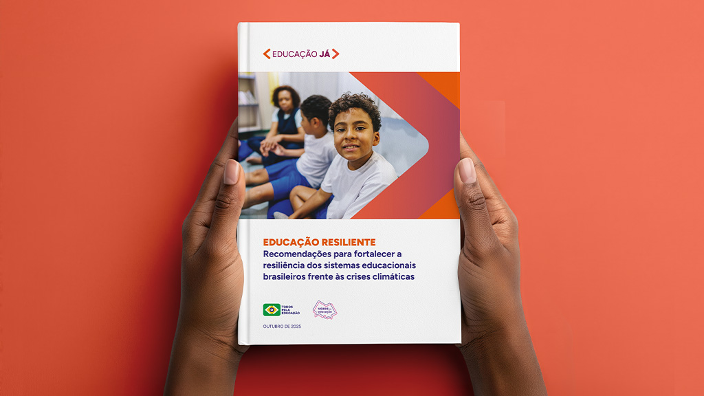 A imagem mostra um par de mãos segurando um relatório impresso com capa branca. Na capa há uma foto de um garoto sorrindo em sala de aula, acompanhada de um gráfico em forma de seta nas cores laranja e roxa. O título do documento é “Educação Resiliente – Recomendações para fortalecer a resiliência dos sistemas educacionais brasileiros frente às crises climáticas”. A publicação é assinada pelo Todos Pela Educação e pelo movimento Vozes da Educação, com data de outubro de 2025.