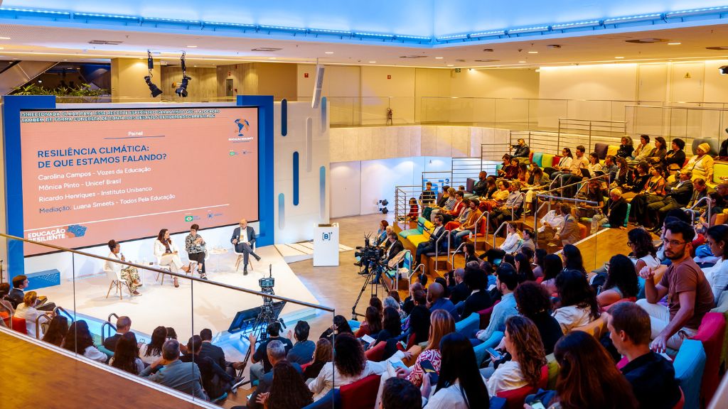 A imagem mostra um auditório moderno e bem iluminado, com público numeroso assistindo a um painel intitulado “Resiliência Climática: de que estamos falando?”. No palco, quatro pessoas estão sentadas em cadeiras transparentes, participando da conversa mediada por Luana Smets, do Todos Pela Educação. O painel conta com Carolina Campos (Vozes da Educação), Mônica Pinto (Unicef Brasil) e Ricardo Henriques (Instituto Unibanco). O ambiente tem arquibancadas coloridas e câmeras posicionadas para registrar o evento. O cenário é profissional e acolhedor, com um grande telão laranja exibindo o nome do painel e dos participantes.