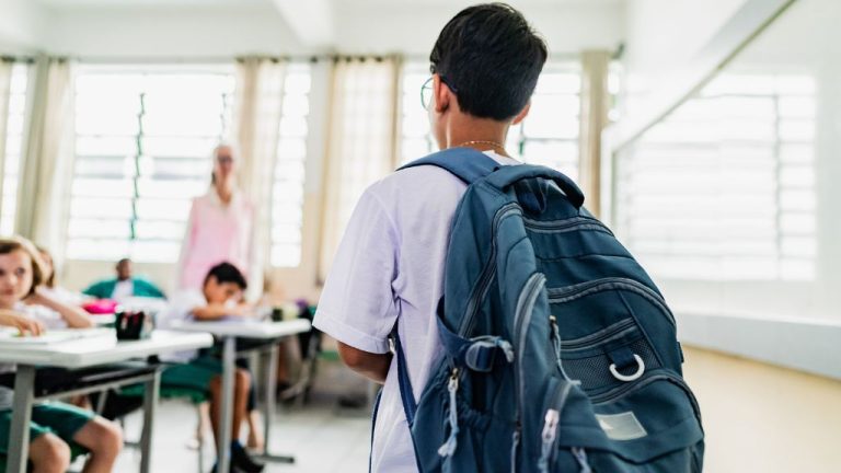 Estudante visto de costas entrando em uma sala de aula, usando mochila azul. À frente, colegas estão sentados em carteiras e uma professora em pé observa a turma.