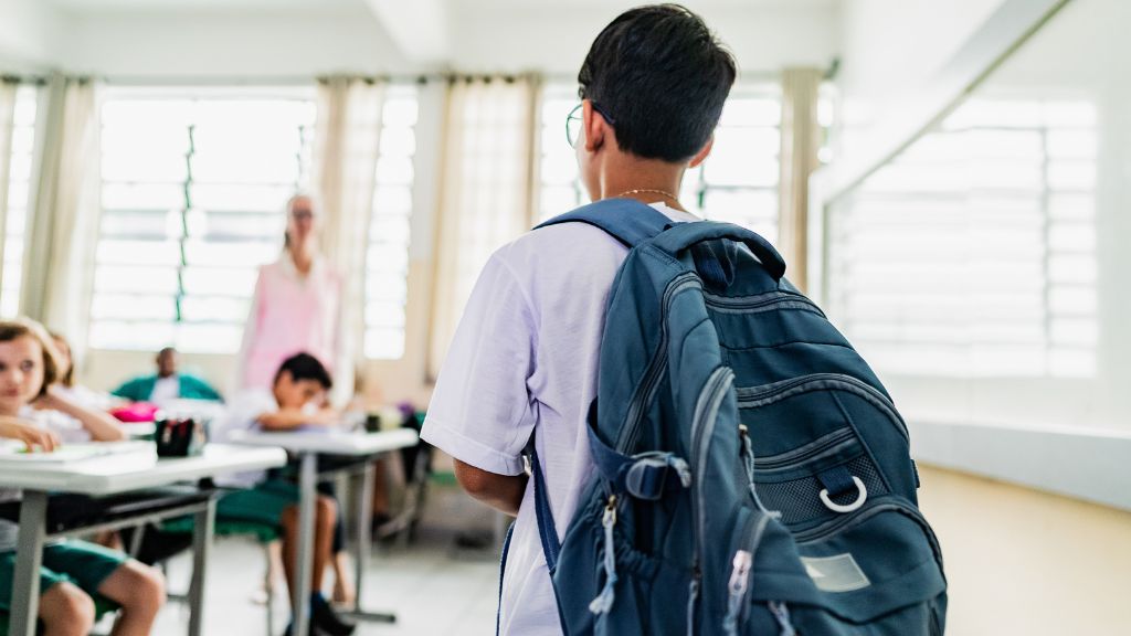 Estudante visto de costas entrando em uma sala de aula, usando mochila azul. À frente, colegas estão sentados em carteiras e uma professora em pé observa a turma.