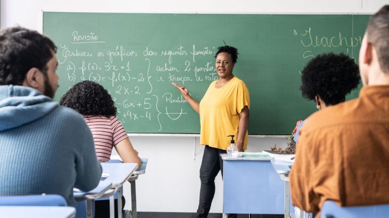 Professora em sala de aula aponta para o quadro verde com anotações de matemática, enquanto estudantes sentados em carteiras observam atentamente. O ambiente é iluminado e organizado, com clima de concentração e aprendizado.
