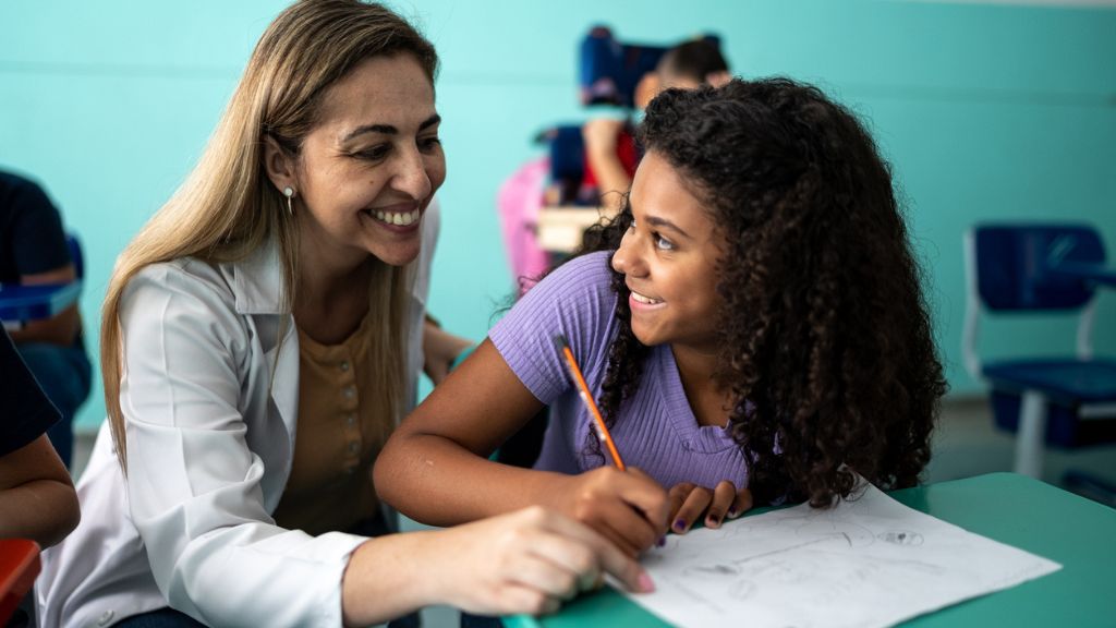 Professora sorri enquanto auxilia uma aluna que desenha em sala de aula. As duas trocam um olhar de cumplicidade e alegria. Ao fundo, outros estudantes estão sentados.
