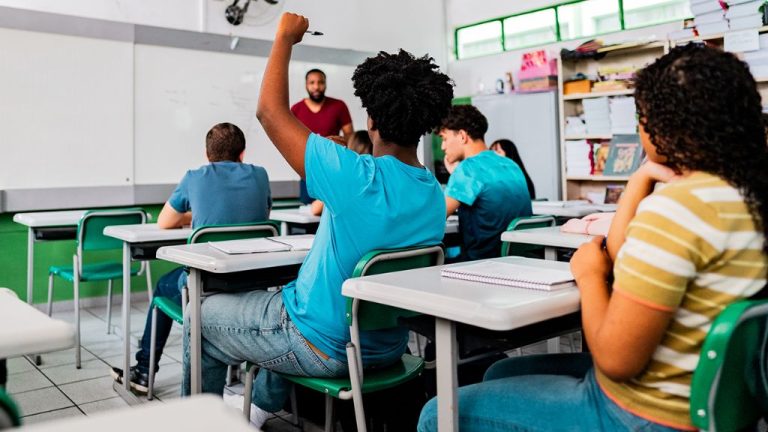 Imagem ilustrativa para o conteúdo sobre o Estudo Conclusão na Educação Básica. A imagem mostra uma sala de aula com estudantes adolescentes sentados em carteiras organizadas em filas. No centro, um estudante de camiseta azul ergue a mão, como se estivesse fazendo uma pergunta ou participando da explicação. Ao fundo, um professor em pé, próximo ao quadro branco, observa a turma enquanto fala. O ambiente é bem iluminado, com janelas altas, paredes brancas e detalhes em verde. Há cadernos e materiais escolares sobre as mesas, e ao lado direito aparece uma estante cheia de livros. A cena transmite um clima de atenção e participação ativa durante a aula.