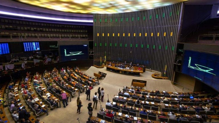 A imagem mostra o plenário da Câmara dos Deputados durante uma sessão, com parlamentares distribuídos em assentos semicirculares, a mesa diretora ao centro e o grande painel verde-amarelo ao fundo. Telões laterais exibem gráficos e há pessoas circulando na área central.
