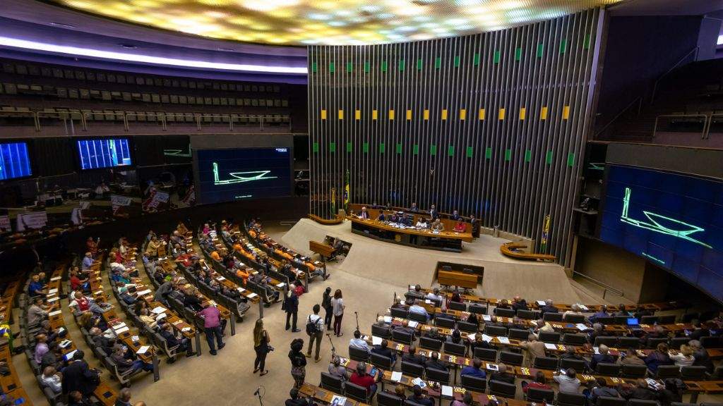 A imagem mostra o plenário da Câmara dos Deputados durante uma sessão, com parlamentares distribuídos em assentos semicirculares, a mesa diretora ao centro e o grande painel verde-amarelo ao fundo. Telões laterais exibem gráficos e há pessoas circulando na área central.