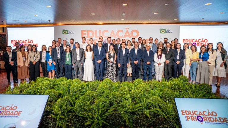 A foto mostra um grande grupo de pessoas posando para a câmera em um auditório, alinhadas em várias fileiras diante de um palco com o letreiro “Educação é Prioridade”. São profissionais e autoridades, muitos usando crachás de evento. O ambiente é iluminado, com painéis e telas exibindo o tema do encontro e uma fileira de plantas decorativas à frente do grupo.