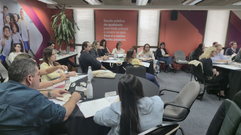Grupo de adultos participa de uma formação em uma sala de eventos, sentados em mesas redondas com materiais de anotação.