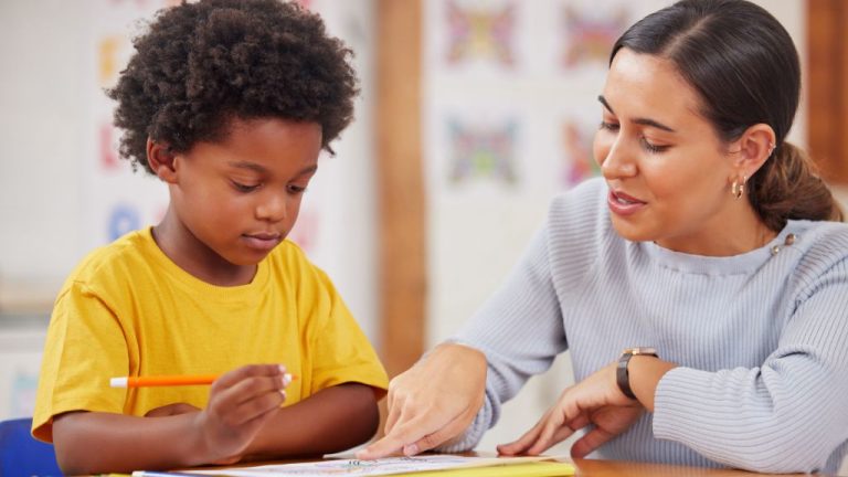 Professora aponta para filha de papel na frente da criança, que olha atento enquanto segura um lápis.