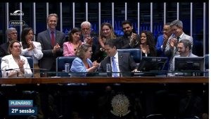 Imagem do plenário do Senado durante uma sessão, com vários parlamentares reunidos. No centro, duas pessoas sentadas trocam um gesto de cumprimento enquanto são aplaudidas pelos demais ao redor, que demonstram satisfação e comemoração. O ambiente é formal, com bancadas, microfones e painéis ao fundo, e a transmissão indica tratar-se da 21ª sessão plenária.