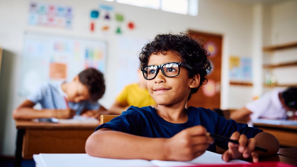 A imagem mostra uma criança com óculos sentada em uma carteira escolar, segurando um lápis e olhando para frente com expressão atenta e confiante. Ao fundo, outros alunos aparecem concentrados em suas atividades, em uma sala de aula bem iluminada e colorida, com cartazes educativos nas paredes.