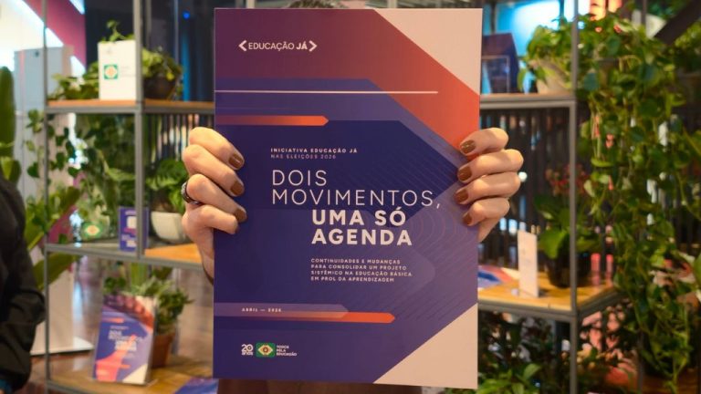 Pessoa segura um folheto com o título “Dois movimentos, uma só agenda”, ligado à iniciativa Educação Já. Ao fundo, há estantes com plantas em um ambiente interno moderno e desfocado.