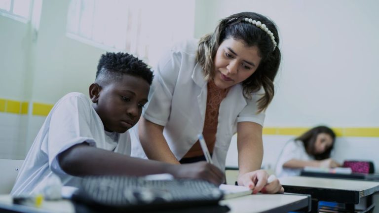 Uma professora auxilia um aluno durante uma atividade em sala de aula. O menino está sentado escrevendo em uma folha, enquanto a professora se inclina ao lado dele apontando para o exercício. Ao fundo, outra estudante aparece desfocada em uma carteira. O ambiente é iluminado, com paredes claras e detalhes amarelos.